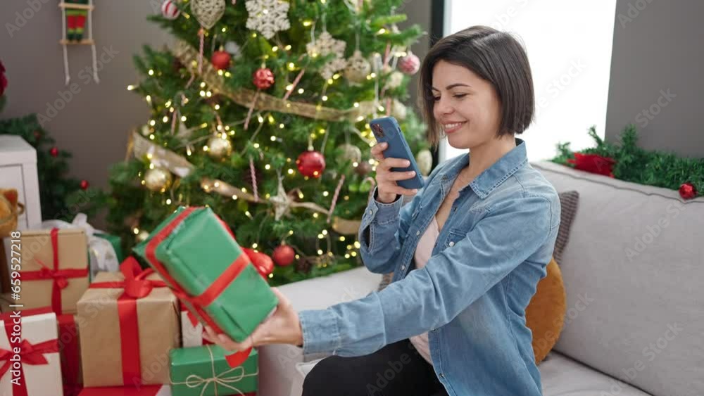 Young caucasian woman taking photo of present sitting by christmas tree at home Young caucasian woman taking photo of present sitting by christmas tree at home