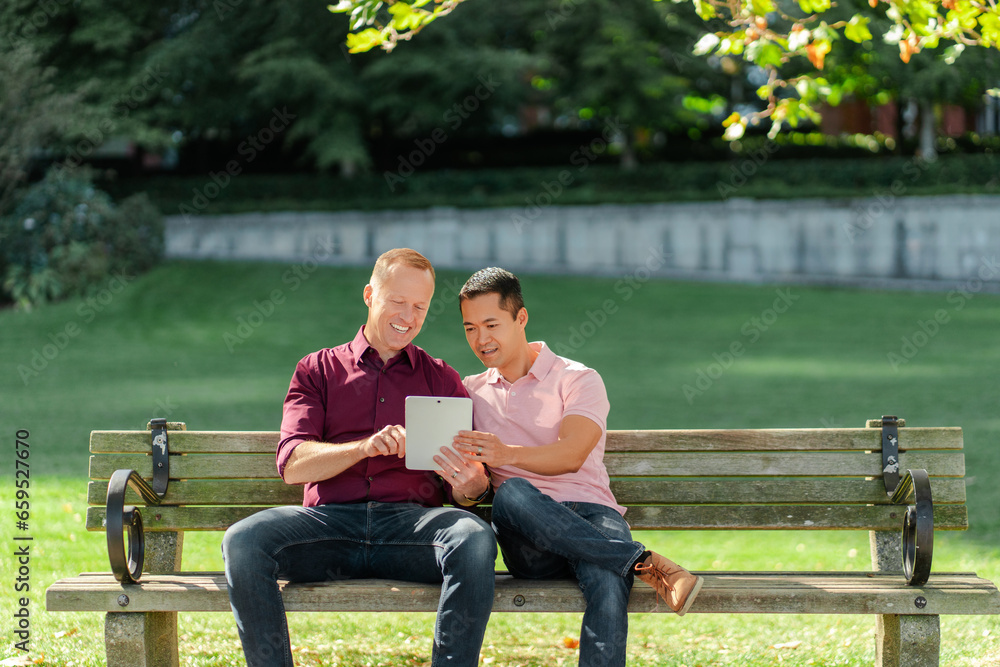 Middle aged positive gay couple, homosexuals sitting on bench on street, using digital tablet Middle aged positive gay couple, homosexuals sitting on bench on street, using digital tablet