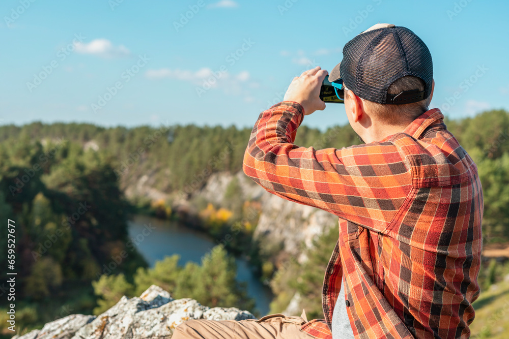 man in cap red plaid shirt sitting in rocks and taking photo on smartphone of mountains autumn forest and river landscape travel and tourism copy space using modern technology