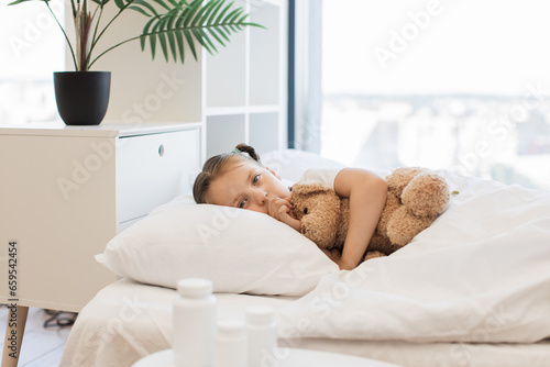 Fatigued cute girl with influenza coughing in clenched fist while lying on bed with soft white linen. Little blond child finding comfort in embrace of her teddy bear toy.