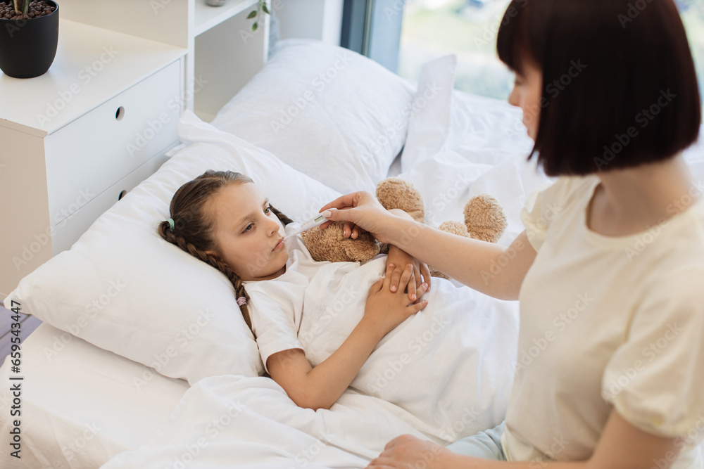 Fototapeta premium Fatigued little girl lying in bed under soft blanket while mother sitting near and checking temperature with electronic thermometer. Concept of diseases and health treatment at home.
