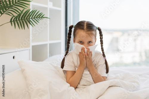 Caucasian ill girl with two braids using white tissue for running nose while sitting on comfy white bed. Tired female child suffering from sneezing during influenza at home.
