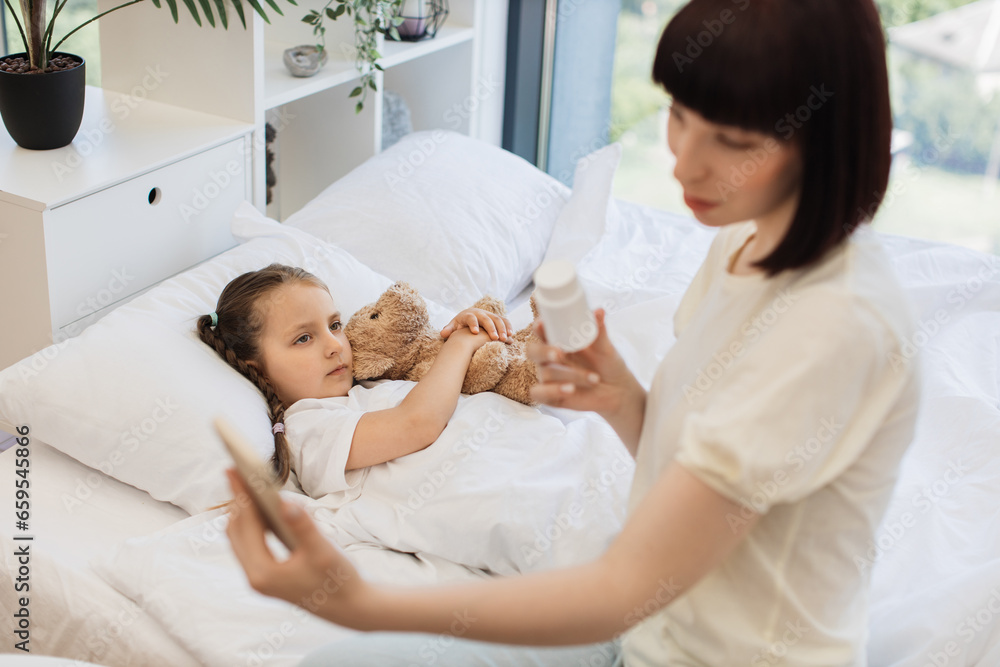 Caring mother communicating with doctor via video call on modern smartphone and showing pills that her sick daughter taking. Cute girl lying in bed and hugging teddy bear while waiting for help. Caring mother communicating with doctor via video call on modern smartphone and showing pills that her sick daughter taking. Cute girl lying in bed and hugging teddy bear while waiting for help.