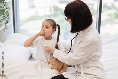 Qualified caucasian nurse listening breathing organs of small adorable child with stethoscope and asking her to cough. Friendly doctor examining little girl for further treatment at cozy bedroom.