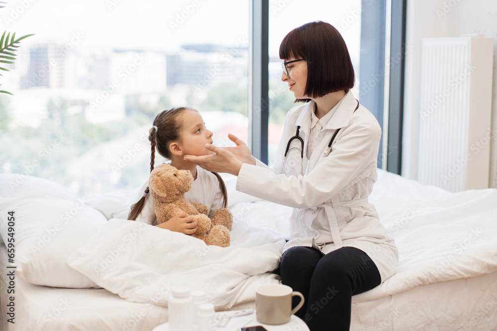 Competent female doctor conducting detailed examination of adorable little girl sitting on cozy bed and hugging teddy bear. Medical worker checking lymph nodes of sick patient during domestic visit. Competent female doctor conducting detailed examination of adorable little girl sitting on cozy bed and hugging teddy bear. Medical worker checking lymph nodes of sick patient during domestic visit.