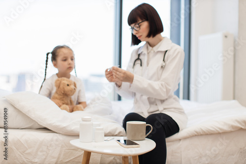 Attractive brunette pediatrician holding electronic thermometer in hands while using to measuring temperature of sick girl. Smiling woman thinking about treatment to prescribing for small child.