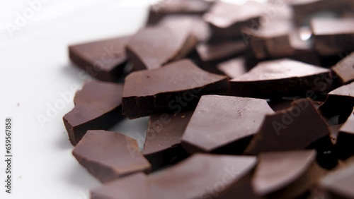 Dark chocolate bars in broken pieces on ceramic plate surface. Baking ingredient close up. Rotation