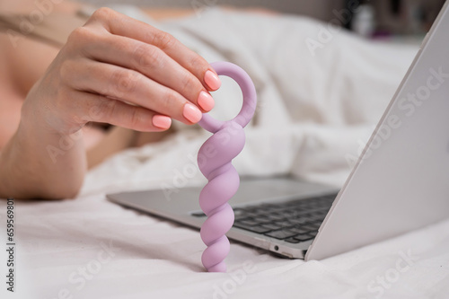 Woman holding lilac anal beads next to laptop while lying on bed. 