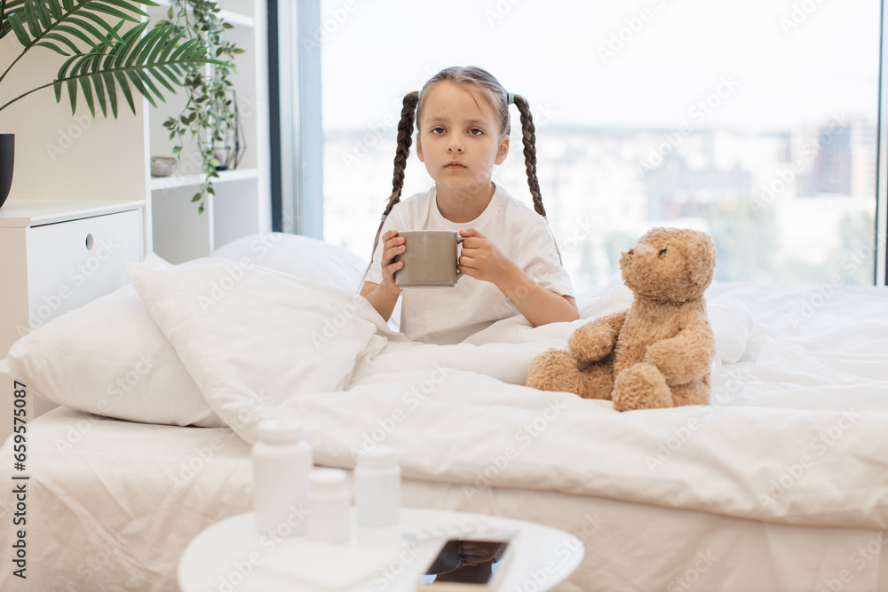 Unhappy caucasian girl resting in cozy bed with cup of hot drink because feeling sore throat indoors. Adorable child trying to swallowing unpalatable pills with delicious tea at home.