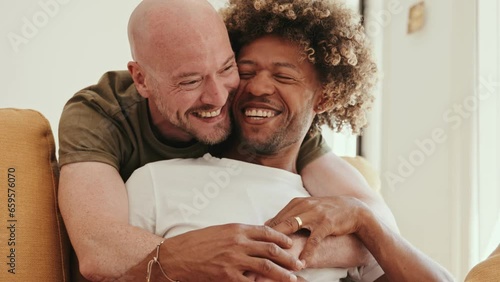 Diverse gay couple at home, sharing love and tenderness on the sofa. A black male with an afro hairstyle smiles, feeling deeply in love with his Caucasian partner, and they share laughter