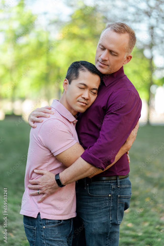 Attractive homosexual family, gay couple hugging with closed eyes standing in park on urban street Attractive homosexual family, gay couple hugging with closed eyes standing in park on urban street