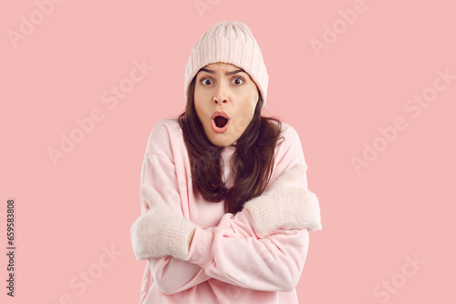 Woman wearing warm light pink hat, sweatshirt and mittens shocked by unexpected extremely low winter temperature feeling very cold, shivering, looking at camera with funny face expression. Studio shot