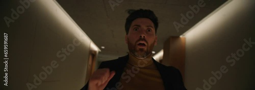 Footage of scared young bearded man in formal clothes running down hallway indoors. Shot of frightened guy in hotel at night. Shot with anamorphic lens. Cinematography technique photo