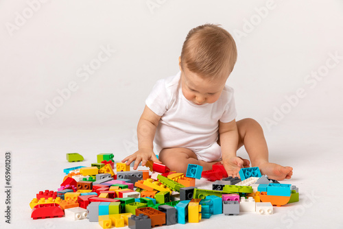 Cute baby boy in white bodysuit playing with a multicolored plastic constructor on a white background isolate, child development concept, space for text
