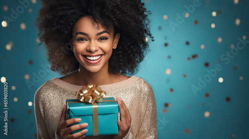 Happy black woman holding a blue gift box for Christmas, isolated on blue background with silver ornament. Xmas party celebration concept