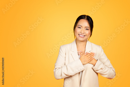 Asian millennial woman places hands on chest, expressing sincere gratitude, orange studio backdrop