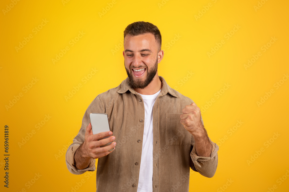 Glad confident european young guy with beard look at smartphone, celebrate win with fist sign Glad confident european young guy with beard look at smartphone, celebrate win with fist sign