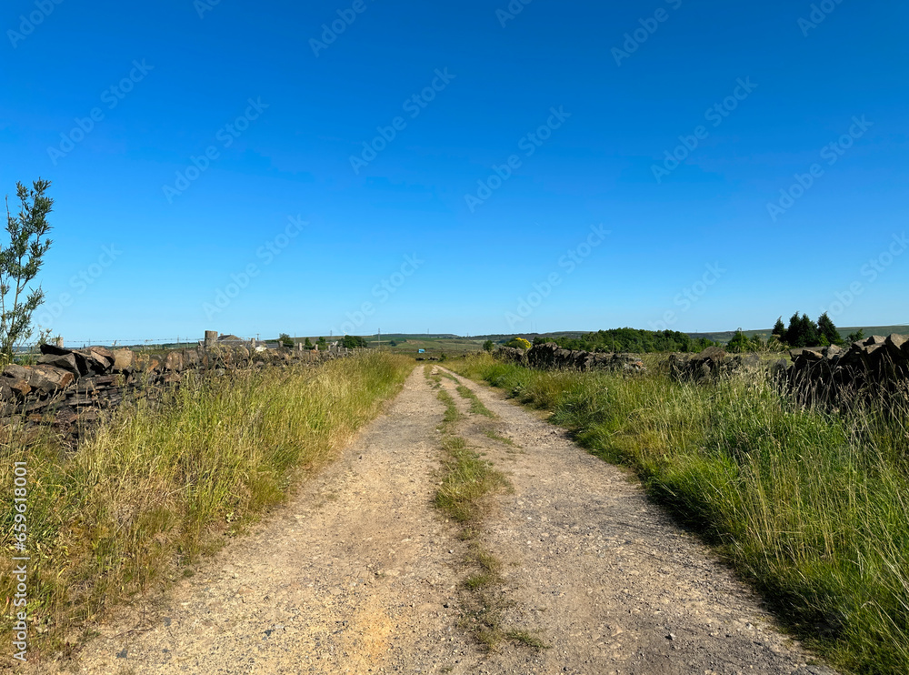 Obraz premium Dirt track, with dry stone walls, wild plants, and grasses, leading past fields, to farms in, Ripponden, UK