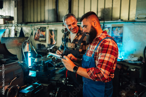 Two metallurgists talking about a metal cylinder next to a lathe machine.