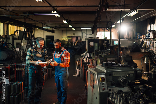 A wide shot of two factory workers in a hangar with various parts and machines.