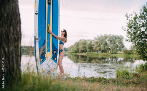 A young woman with an open swimsuit stands with a sapboard on the shore of the lake. Stand Up Paddle. SUP touring photo