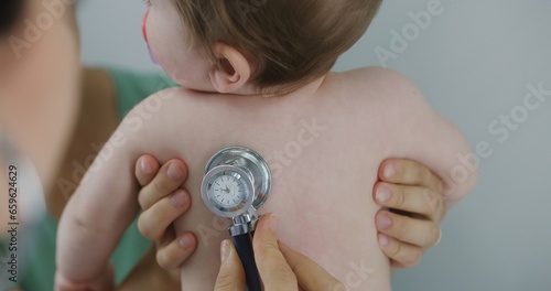 Fototapeta Naklejka Na Ścianę i Meble -  Happy mother holds on hands her handsome baby with pacifier in mouth. Pediatrician uses stethoscope to listen to heartbeat and lungs of cute little child. Physician does checkup in hospital. Close up.