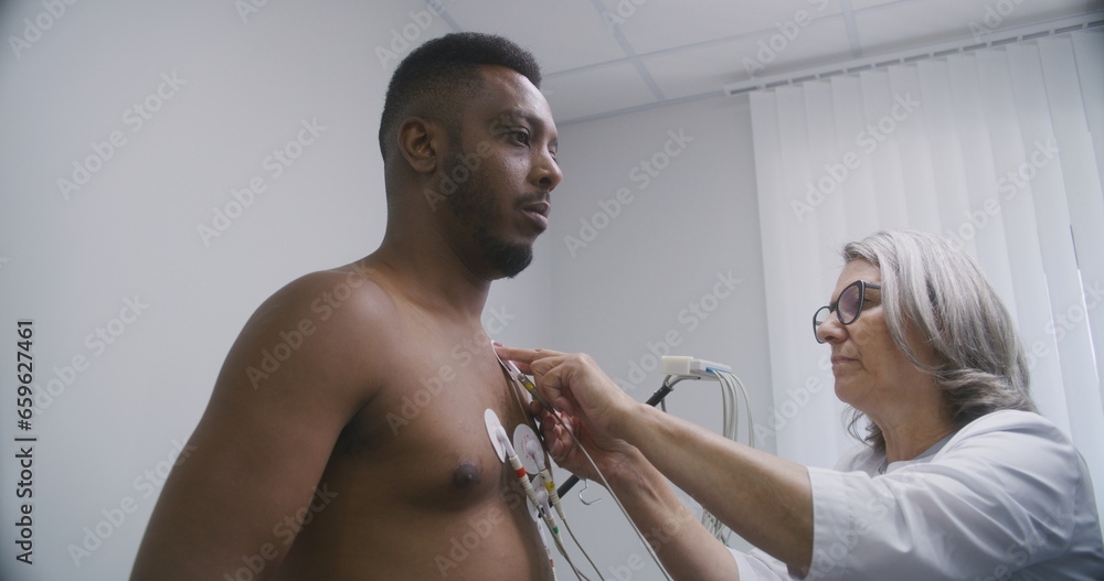 Female cardiologist, doctor puts holter sensors on patient with cardiovascular disease. African American man during functional rehabilitation in cardiology room. Electrocardiography in modern hospital Female cardiologist, doctor puts holter sensors on patient with cardiovascular disease. African American man during functional rehabilitation in cardiology room. Electrocardiography in modern hospital