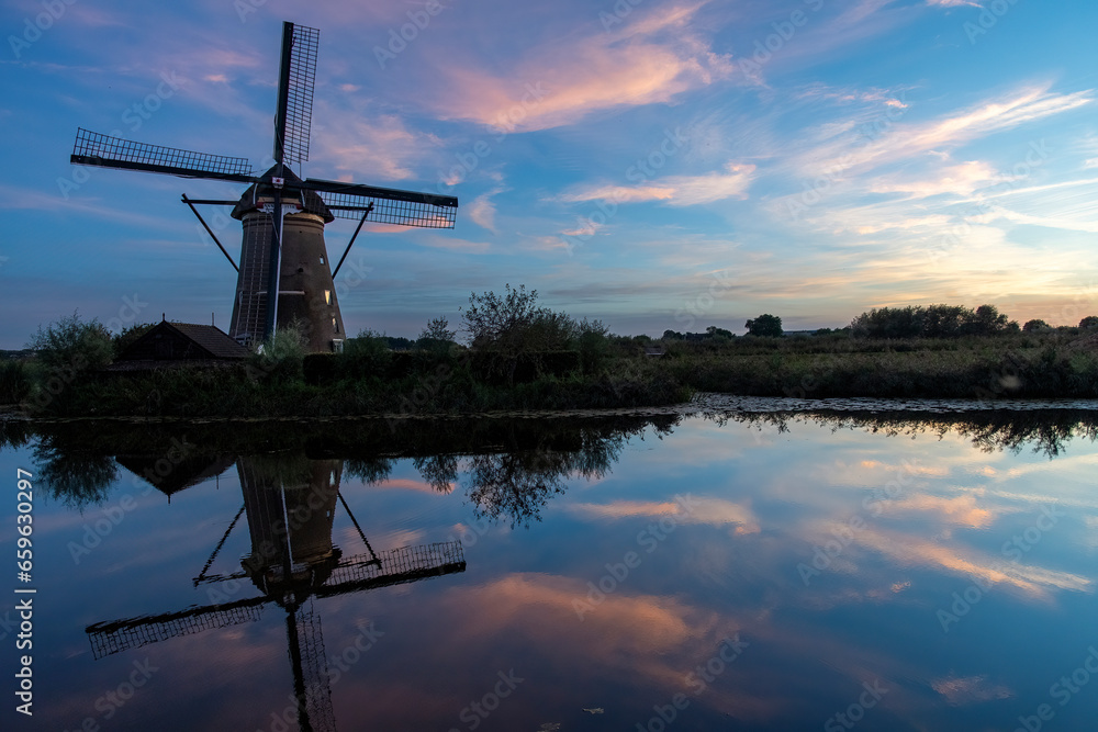 Sunset with typical Dutch windmill along long row of reed beds in the Kinderdijk area, the Netherlands with perfect reflection of windmill, reed beds and coloring cloudscape in tranquil water