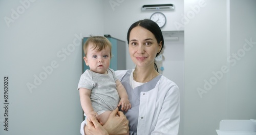 Female pediatrician stands in middle of bright hospital room and looks at camera. Adult doctors holds baby. Little boy looks at camera and holds hand of medic. Medical staff at work in modern clinic. © Framestock