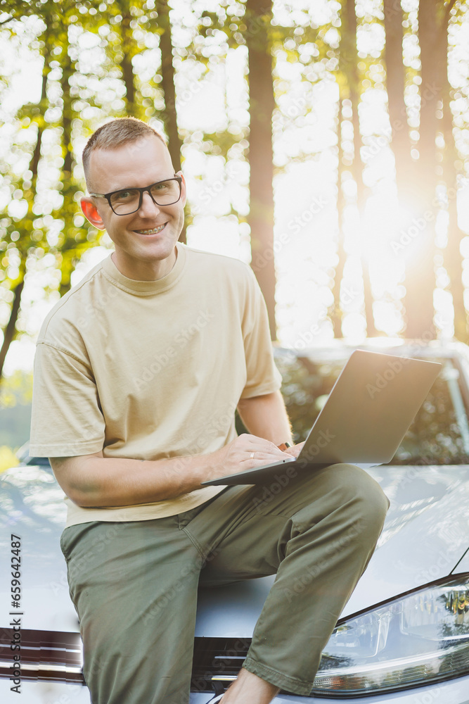 Fototapeta premium A serious man in glasses is working on a laptop on the hood of his car. Remote work in nature. Work on a laptop online. Working on a computer while traveling.