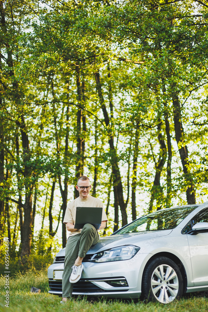 A serious man in glasses is working on a laptop on the hood of his car. Remote work in nature. Work on a laptop online. Working on a computer while traveling. A serious man in glasses is working on a laptop on the hood of his car. Remote work in nature. Work on a laptop online. Working on a computer while traveling.