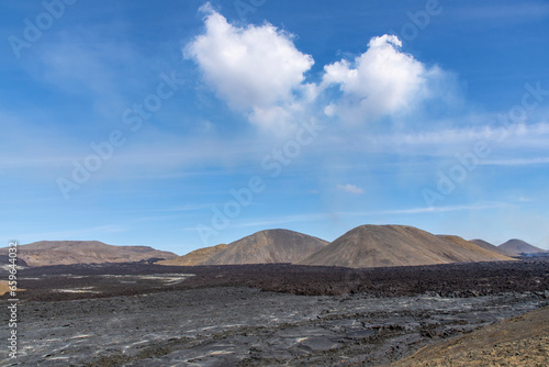 Panoramic view over large black lava field with mountains in volcanic landscape on Reykjanes Peninsula, Iceland, near mountain Fagradalsfjall volcano area against a white clouded blue sky