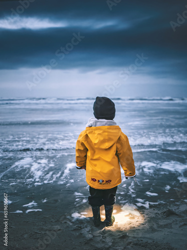 A little boy from behind looks at the horizon in a yellow raincoat. Nature, exterior at night between a stormy sky and a rough sea. Childhood concept. Universal Children's Day.