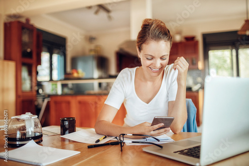 Happy young woman looking at a smartphone while going over paperwork in the kitchen