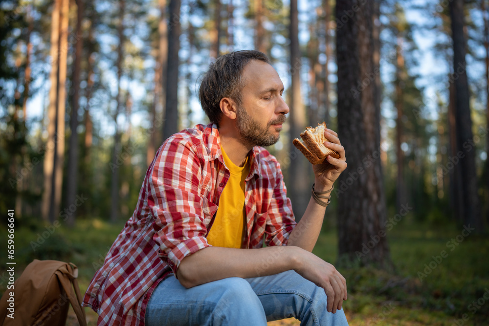 Fototapeta premium Carefree man eating sandwich snack during hike walk travel camping vacation, feels calm relax in nature landscape. Happy hiking male resting taking break with healthy food in forest. Autumn nature.