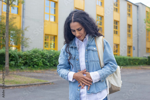 Worried Eastern girl leaves a hospital or school, university, holds her stomach, looks down, writhes in pain, experiences severe discomfort and spasm in the lower abdomen, in casual clothes outdoors.