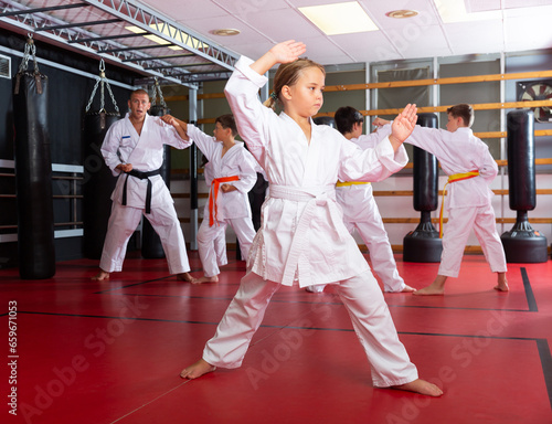 Adolescent girl in kimono posing in gym while other kids sparring in background during karate group training.