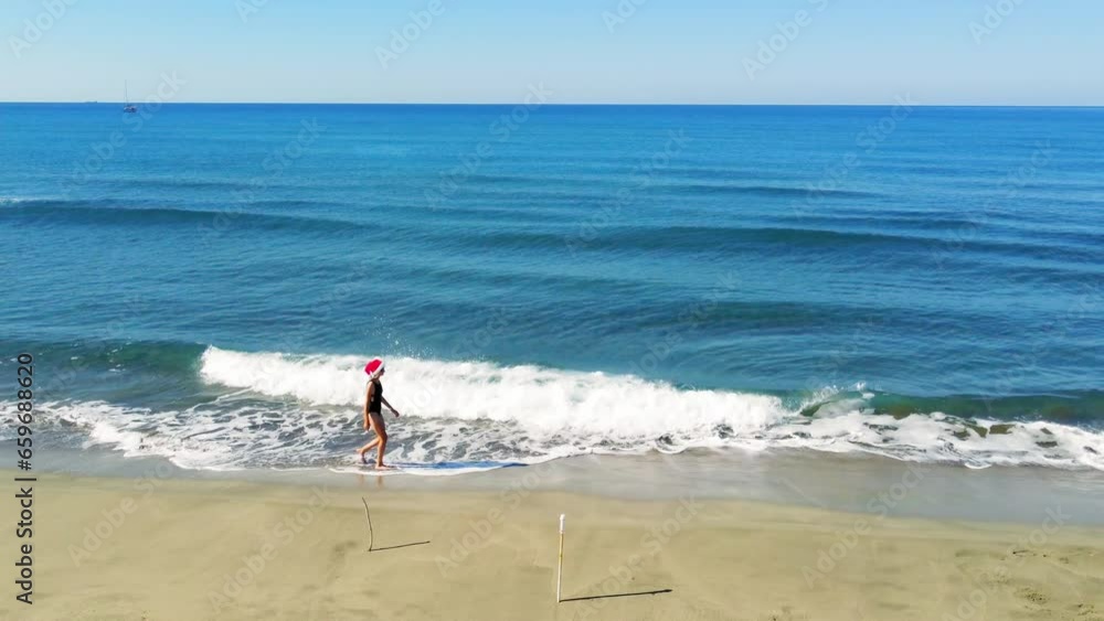 Young beautiful slender woman in Santa Claus hat walking on sandy beach, rejoicing in Christmas or New Year. Shooting with drone. Weekend Sea or Ocean. Resort Vacation. Young beautiful slender woman in Santa Claus hat walking on sandy beach, rejoicing in Christmas or New Year. Shooting with drone. Weekend Sea or Ocean. Resort Vacation.