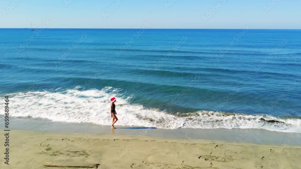 Young beautiful slender woman in Santa Claus hat walking on sandy beach, rejoicing in Christmas or New Year. Shooting with drone. Weekend Sea or Ocean. Resort Vacation. Young beautiful slender woman in Santa Claus hat walking on sandy beach, rejoicing in Christmas or New Year. Shooting with drone. Weekend Sea or Ocean. Resort Vacation.
