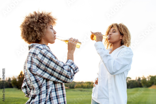 two african american curly women talking and holding bottles of beer in the park at sunset