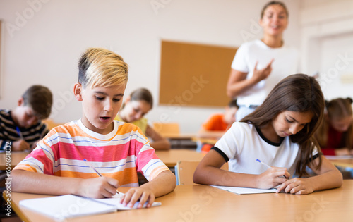 Diligent elementary school student tween boy studying with classmates, making notes of teacher lecture