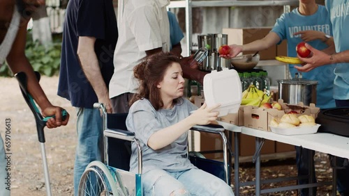 Helpful guy volunteer aids and provides free meal to poor needy woman in wheelchair. Charity workers at outdoor food bank provide humanitarian aid to the crippled and underprivileged individuals.