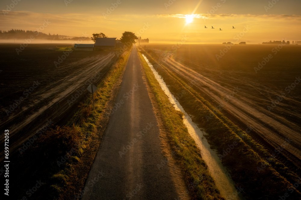 Aerial view of a beautiful sunrise on a rural farmland road. Skagit Valley is the agricultural hub of western Washington state. Historic barns, irrigation canals and plowed fields dominate the area. 