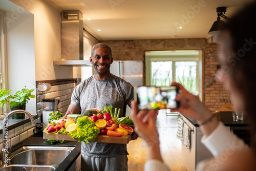 Young diverse couple taking pictures of organic and healthy vegetables in the kitchen at home photo