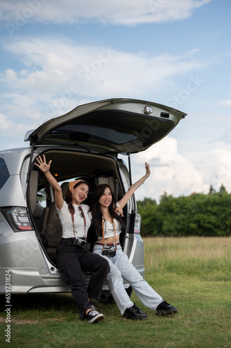 Two cheerful and beautiful Asian female friends are sitting in the car's open trunk.