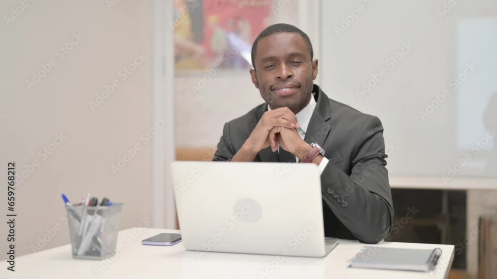 African Businessman Smiling at Camera while Working on Laptop in Office African Businessman Smiling at Camera while Working on Laptop in Office