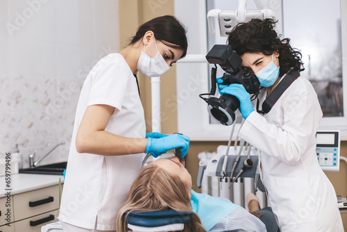 Female dentist with camera making shots of patient s smile after treatment using special shadowless camera with flash ring. Medical assistant holding mirror.