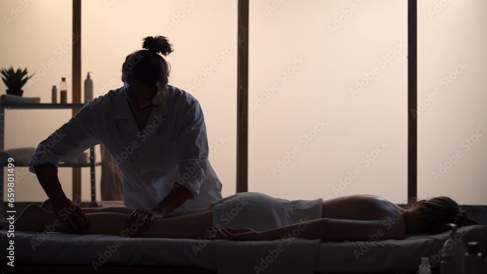 Masseur, massage specialist giving full-body massage to his patient, kneading her legs. Silhouettes of a woman and a man in the massaging room, spa procedure.