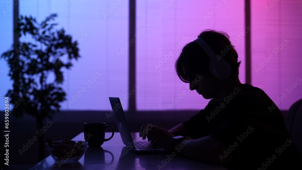 Silhouette close up of male sitting at the food bar alone in neon light, wearing headphones, typing text on laptop.