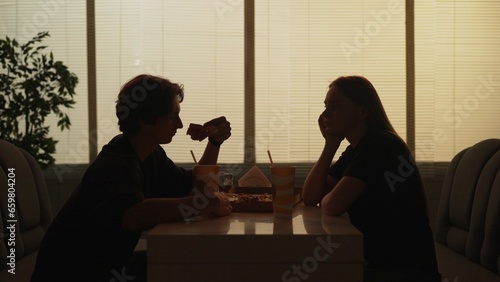 Silhouette of man and girl on a date sitting at the bistro bar together, male eats pizza slice female looking thinking.